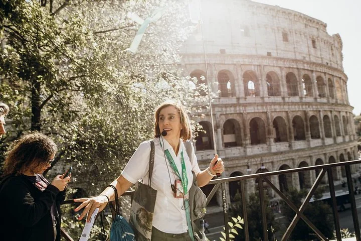 A guide talks beside the Colosseum with sunshine filtering through trees.
