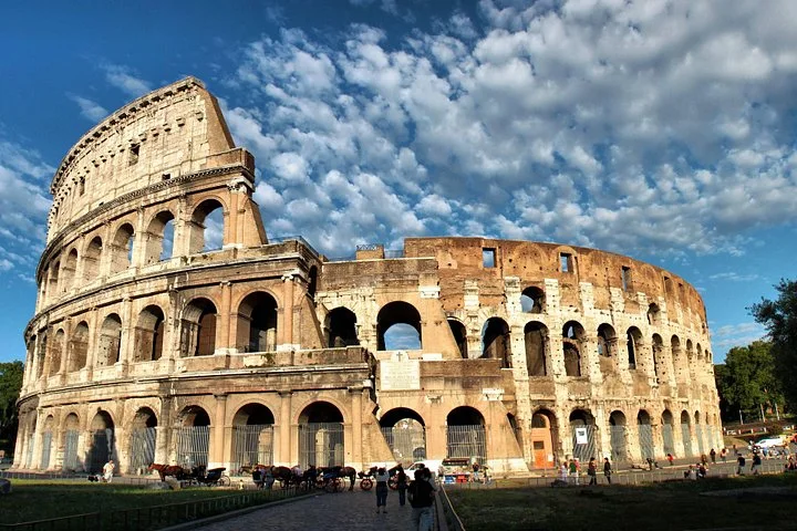 The Colosseum's outer arches stand beneath a bright cloudy sky.
