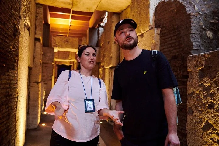 A guide speaks with a visitor among stone pillars below the Colosseum arena.