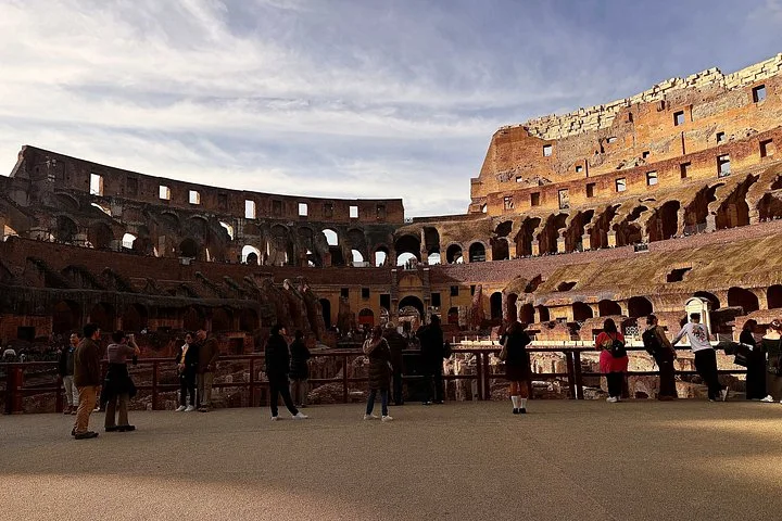 Visitors stand along the Colosseum arena railing in warm light.