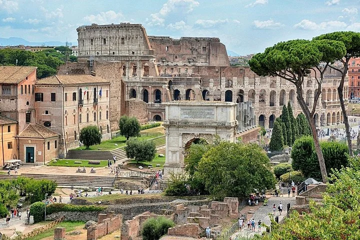 The Arch of Titus and Colosseum rise beyond green ancient ruins.